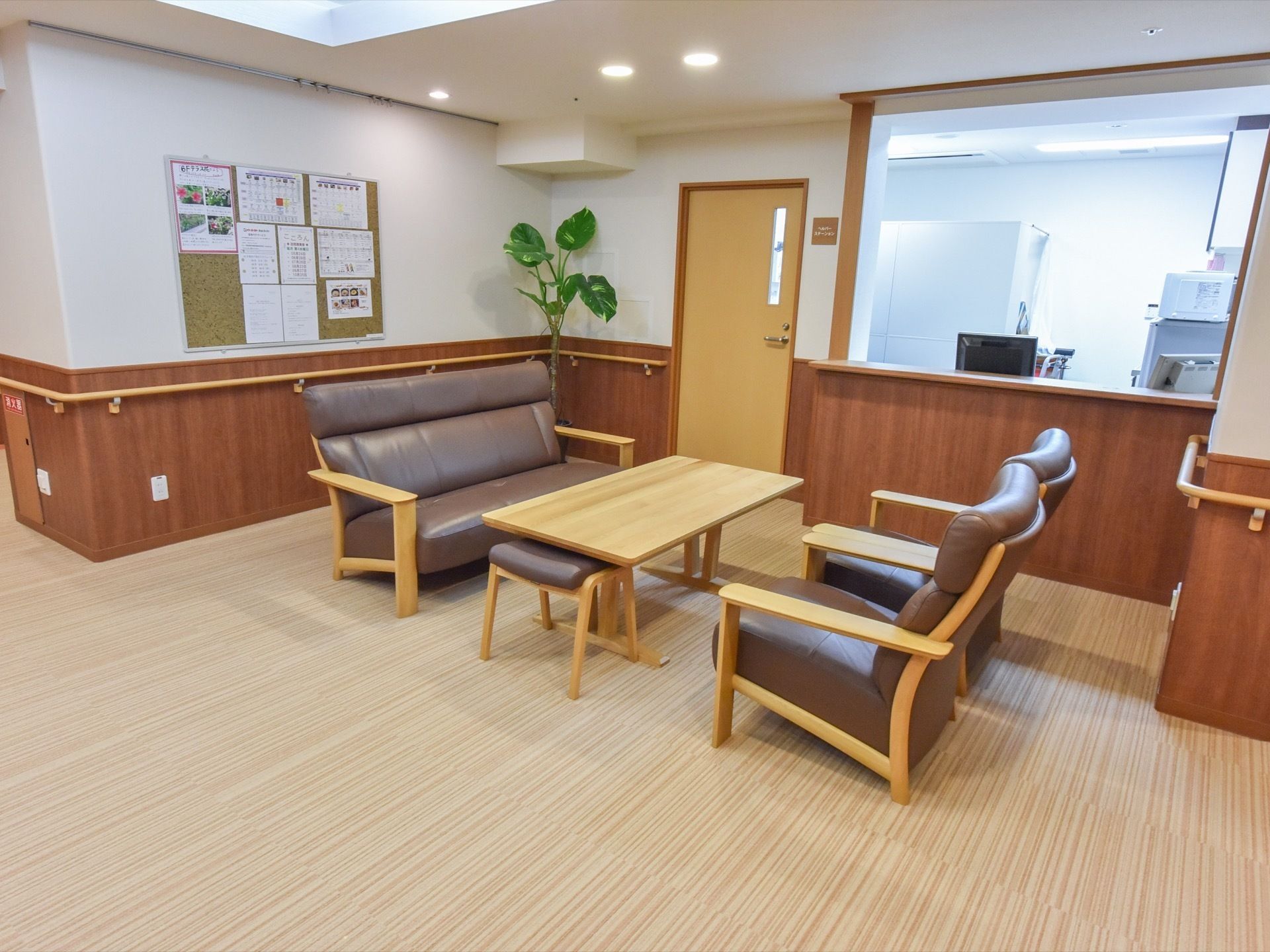 長い手すりが、共有スペース周囲の壁に取り付けられている。壁際には観葉植物が置いてある。奥にはスタッフの部屋がある。