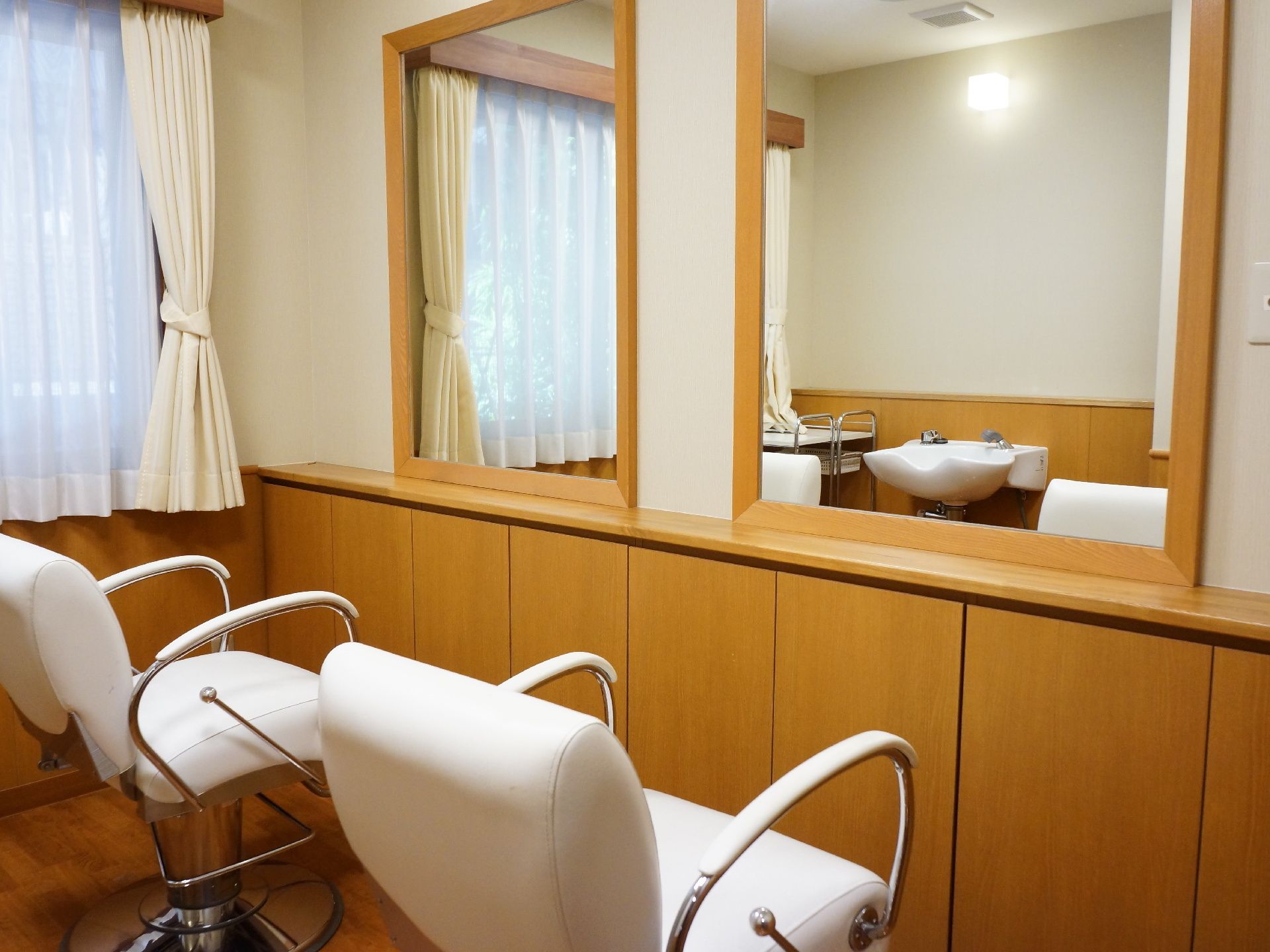散髪用椅子が2つ並んでいる。前にはそれぞれ鏡が付いている。洗髪台は後ろにある。床はフローリングで窓にはカーテンとレースが掛かっている。
