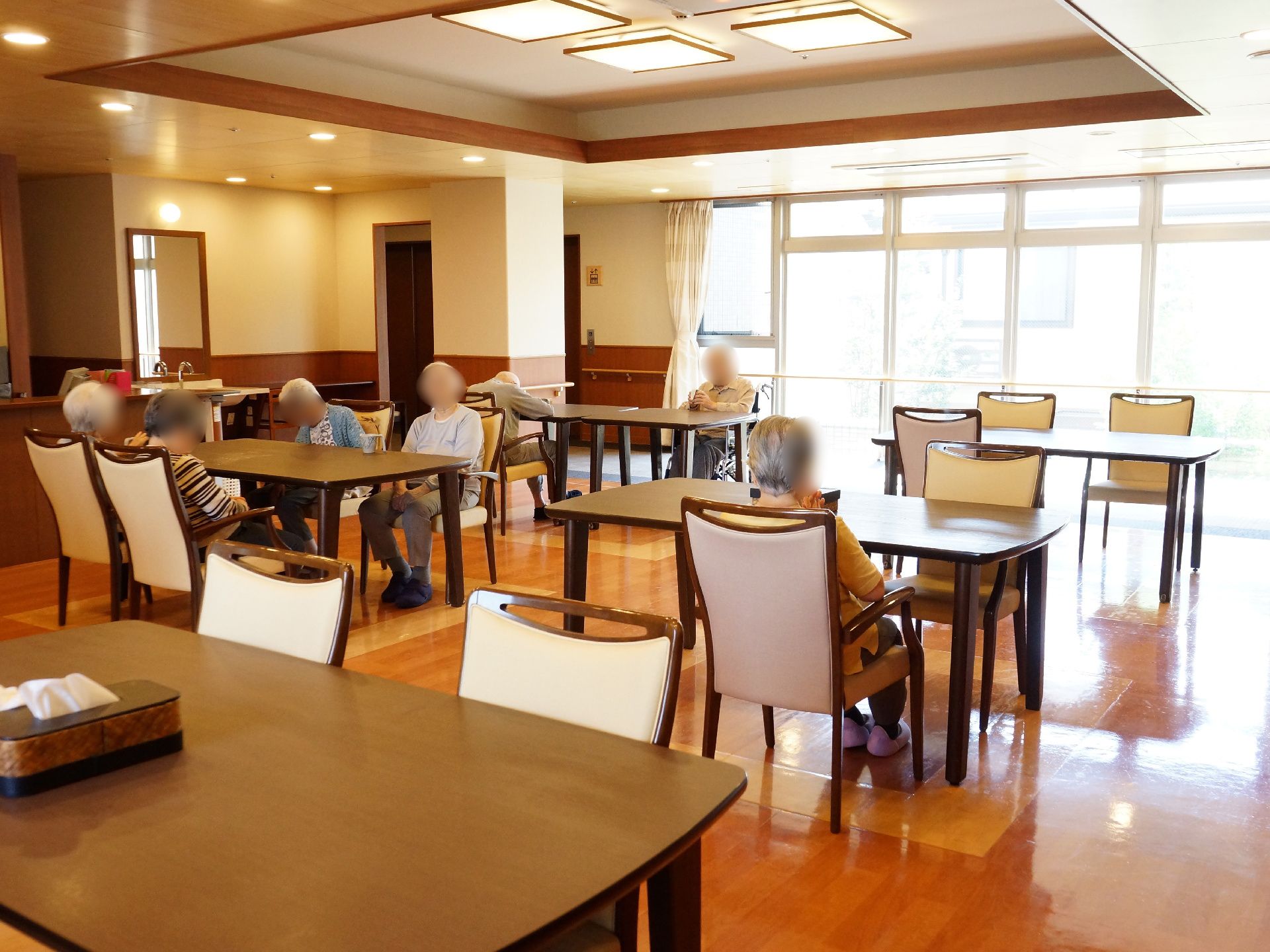 大きな窓ガラスから陽が差し込んでいる部屋にテーブルと椅子がいくつもある。入居者様が椅子でくつろいでいる。