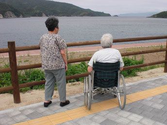 車いすの入居者が女性と一緒に海沿いの道から海を眺めている様子。