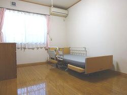 エアコンが設置されたフローリングの部屋には介護ベッドやカーテン付きの窓、タンスなどが置かれている。