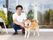 １人の人物と赤い散歩ひもを付けられた１匹の柴犬がいる。その後ろにはホームの玄関とウェルカムボードなどあるのが見える。