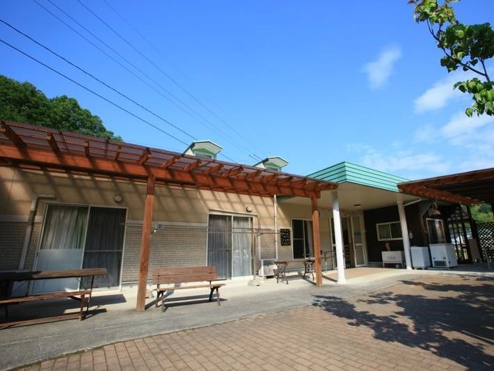 緑に囲まれた立地。平屋建ての建物の玄関の横にはベンチやテーブルが置かれている。