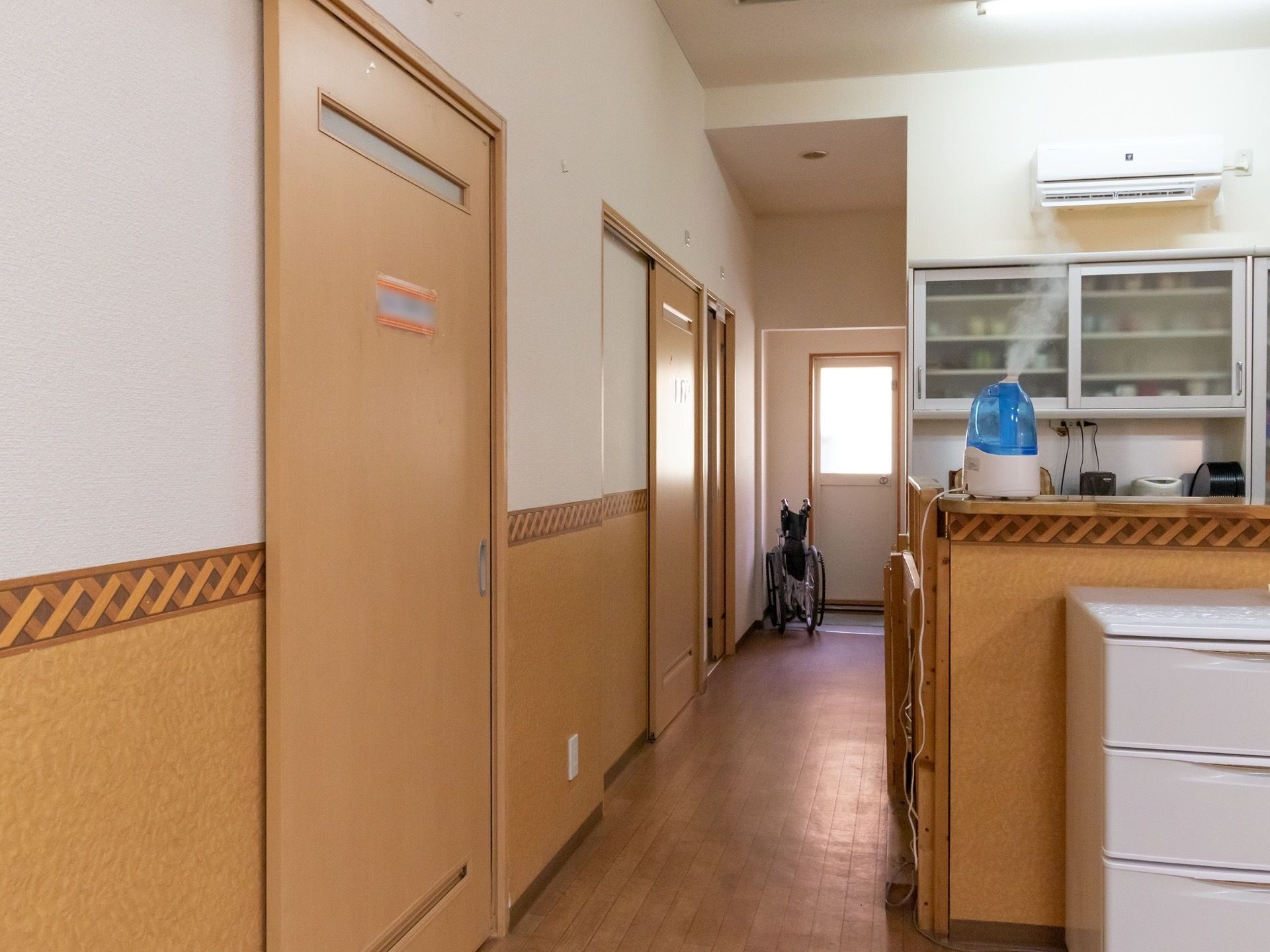 居室が立ち並ぶ廊下で、前にカウンターキッチンが設けられている。突き当りにはドアがあり、車椅子がある。