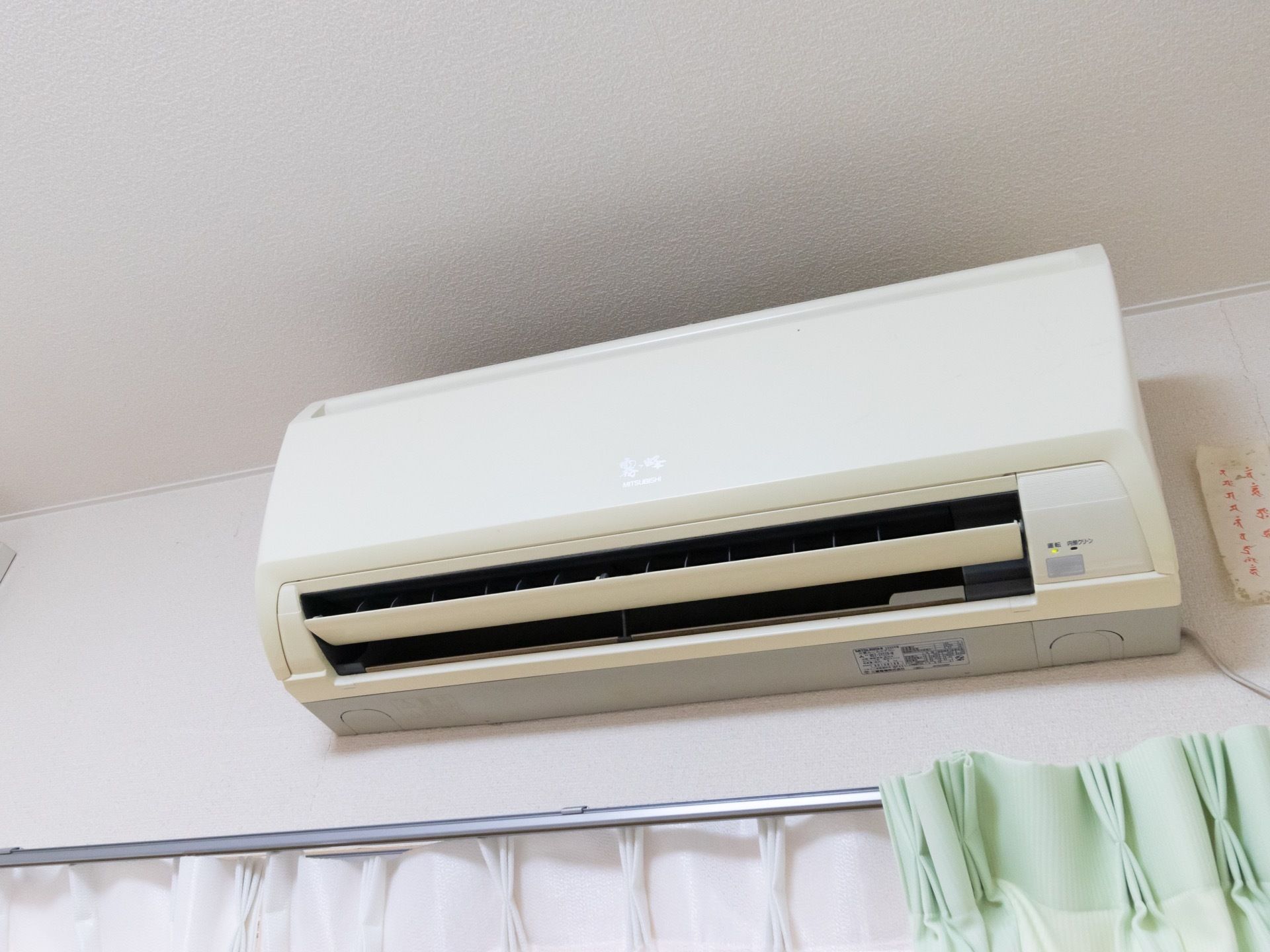 白を基調とした清潔感あふれる明るい部屋。エアコンが１台設置されており、下の窓にはカーテンが取り付けられている。