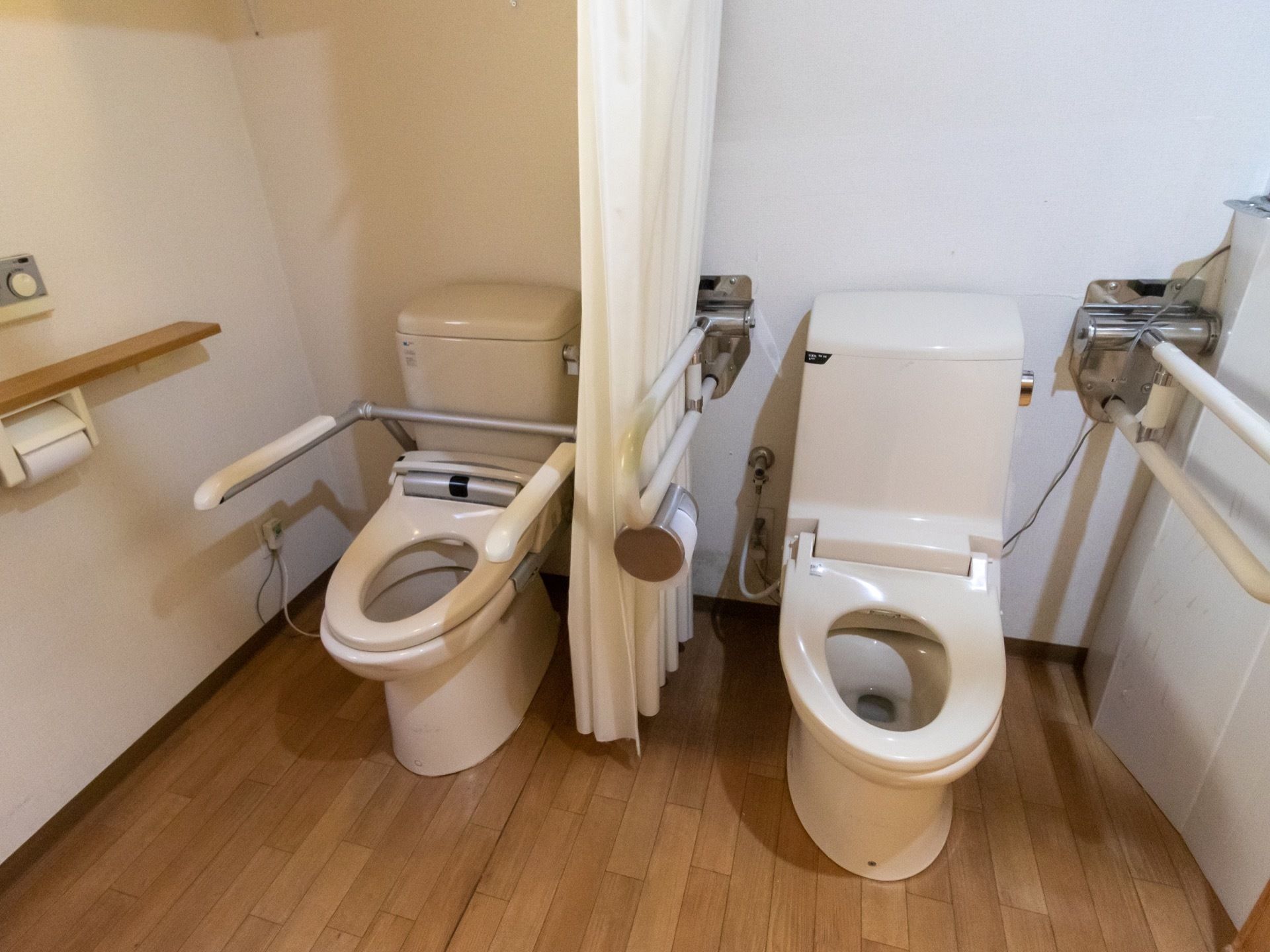 両サイドに手すりが設置されている洋式トイレが２つ並んで設置されている。間は白いカーテンで仕切られている。