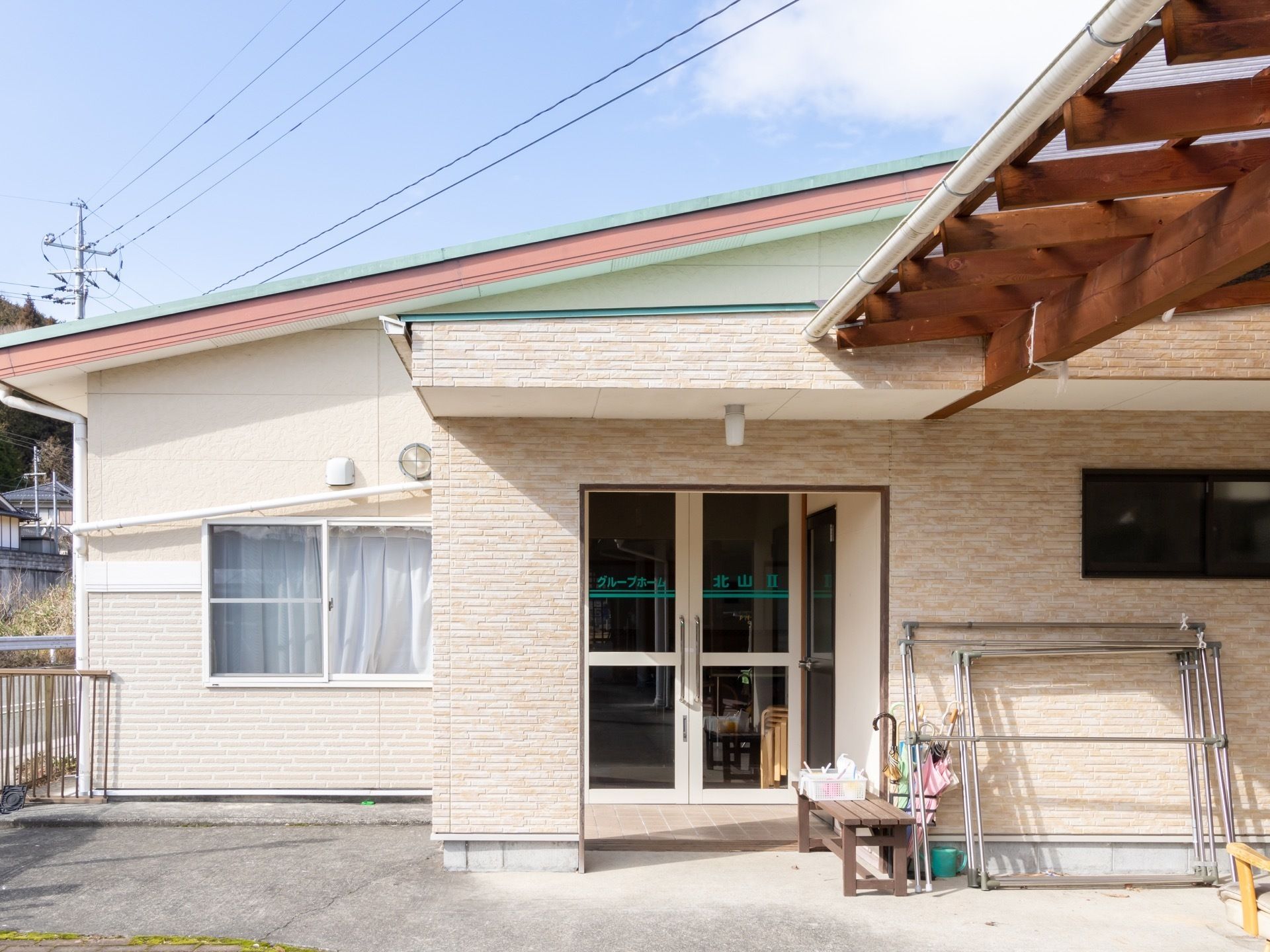 平屋建てのグループホームで、ガラス戸には施設名が書かれている。玄関前には、ベンチや傘立てが用意されている。