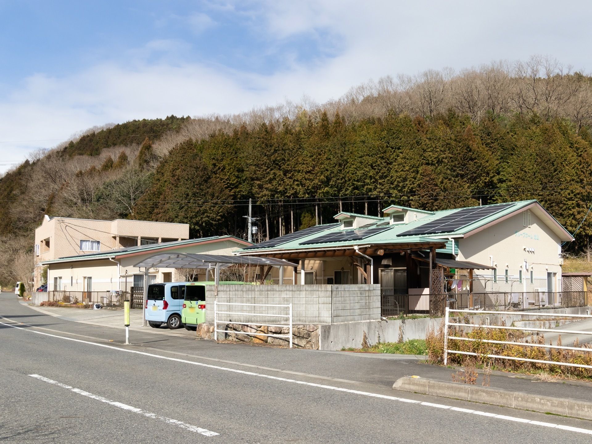 道路に面した場所にある、平屋建ての建物である。建物の前には車が２台停まっており、建物の奥には、森が見える。