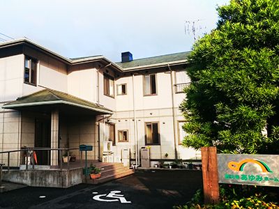 2階建ての建物は白い外壁に緑の屋根の家庭的な雰囲気。玄関横には階段とスロープがある。