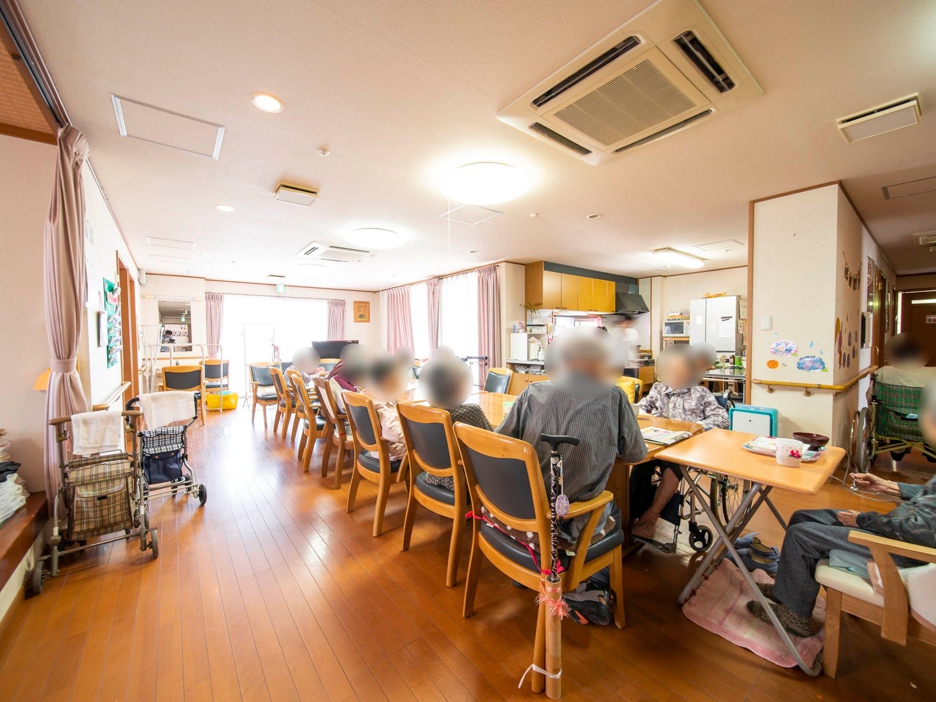 机を縦一列に並べ、入居者様が向かい合わせに座っている。キッチンが併設されており、近くに冷蔵庫が置いてある。