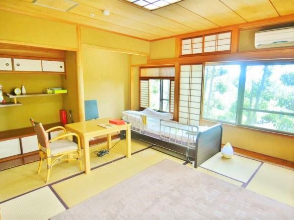 木製の椅子と机などの家具で統一された和風な居室。窓は広く、緑色の木々が見える