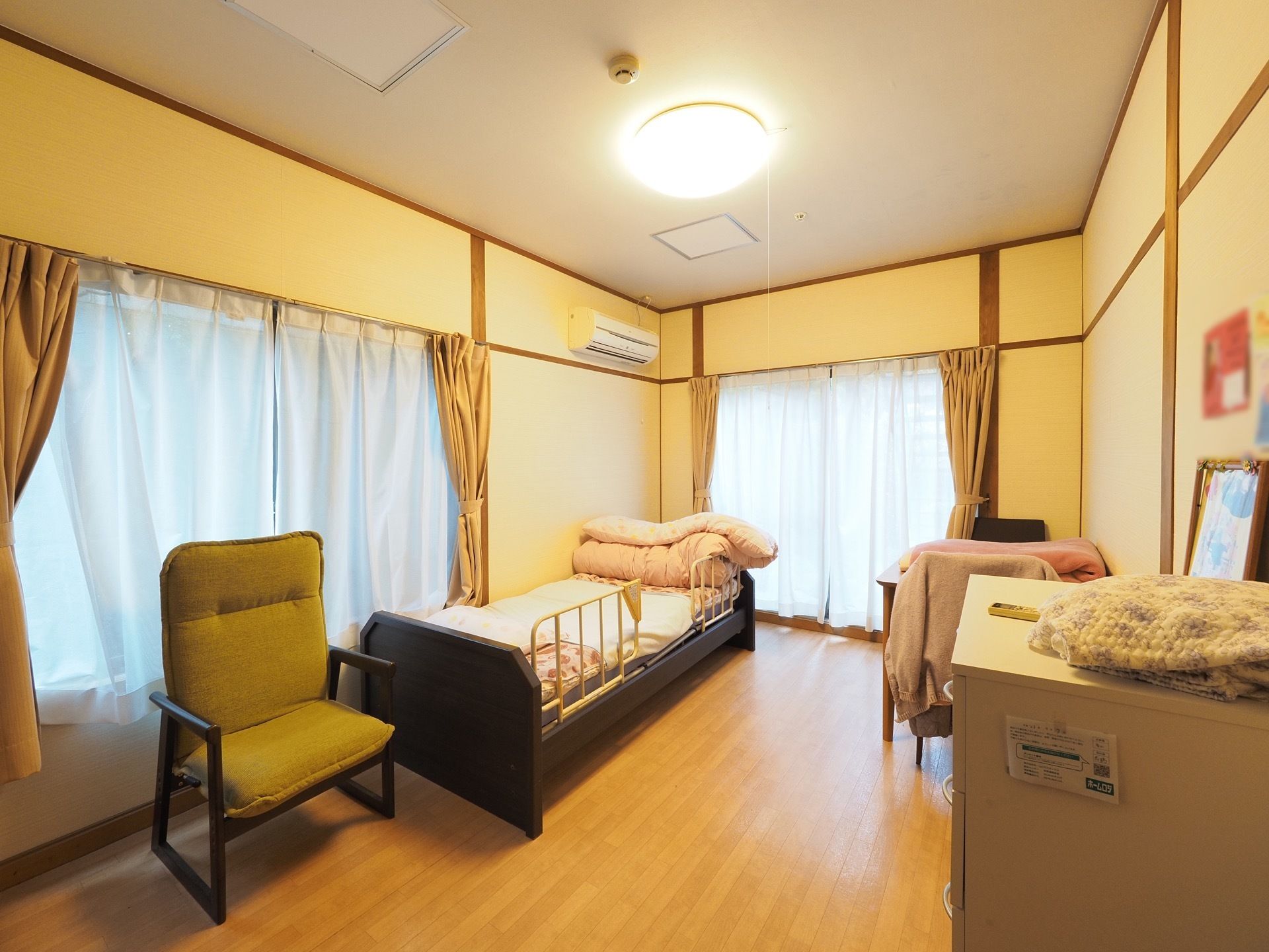 フローリングで、部屋の隅に介護用ベッドとテーブルと椅子が用意されている。大きな窓が２か所用意されており、部屋全体が明るい。