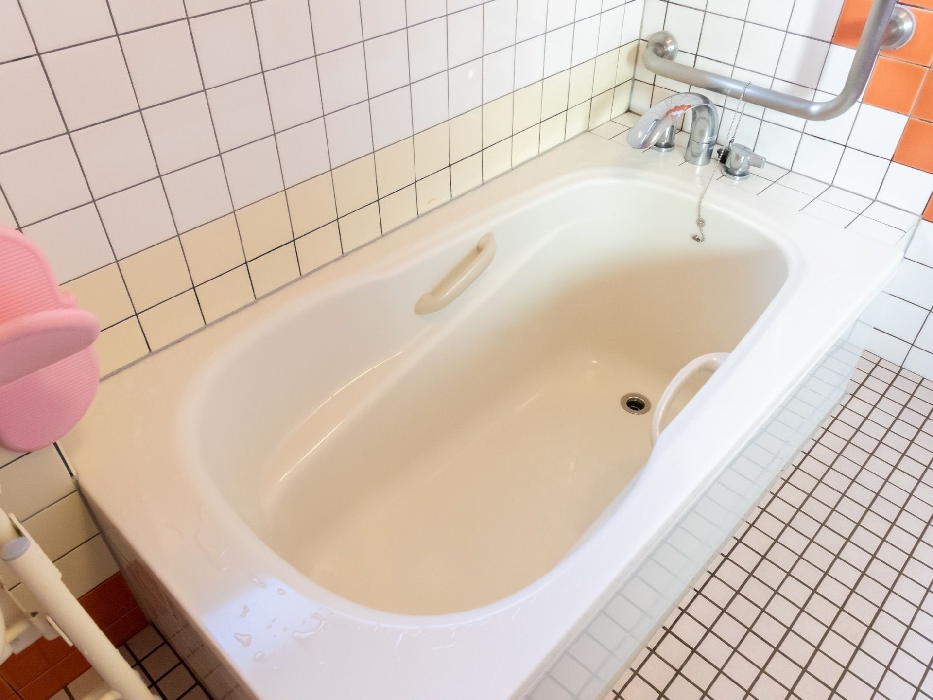 オレンジと白のタイル貼りの浴室の角に手すり付きの浴槽が備え付けられている。浴槽の横の壁にはＬ字型の手すりもある。
