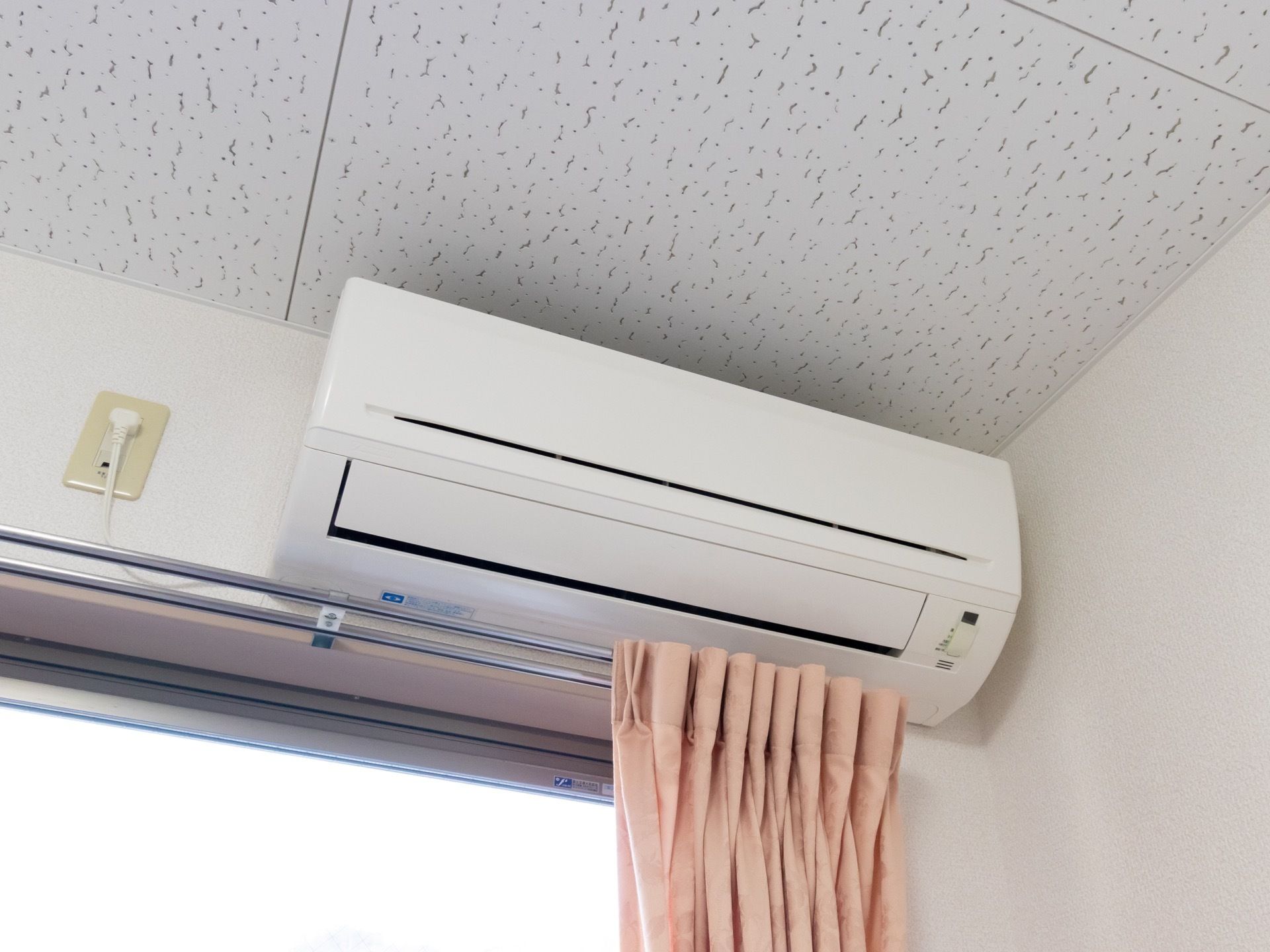 白い壁の部屋に桃色のカーテンのかかった窓がある。窓の上には白いエアコンが１台設置されているのが見える。