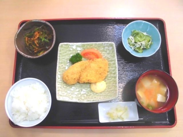 フライをメインに白いご飯やみそ汁、野菜の和え物やヒジキなどが並んだ黒いトレー。