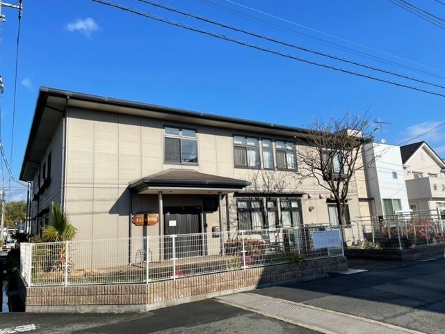フェンスに囲まれた敷地内にはグレーの外壁の２階建ての建物があり、建物の周りには草木が植えられている。