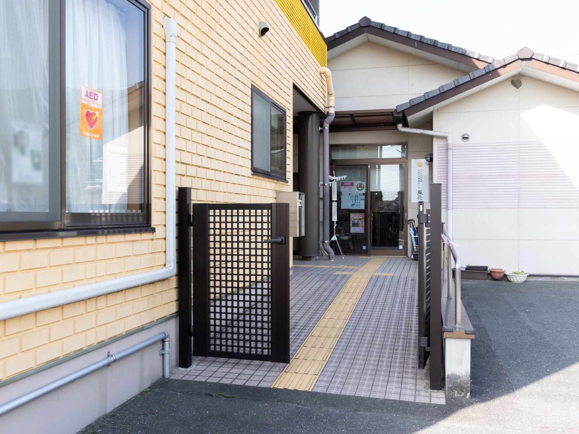 金属製の門扉があり、その横の壁にある窓には、AEDと書かれたステッカーが貼られている。門扉の奥には、玄関へと続くスロープがあるのが見える。