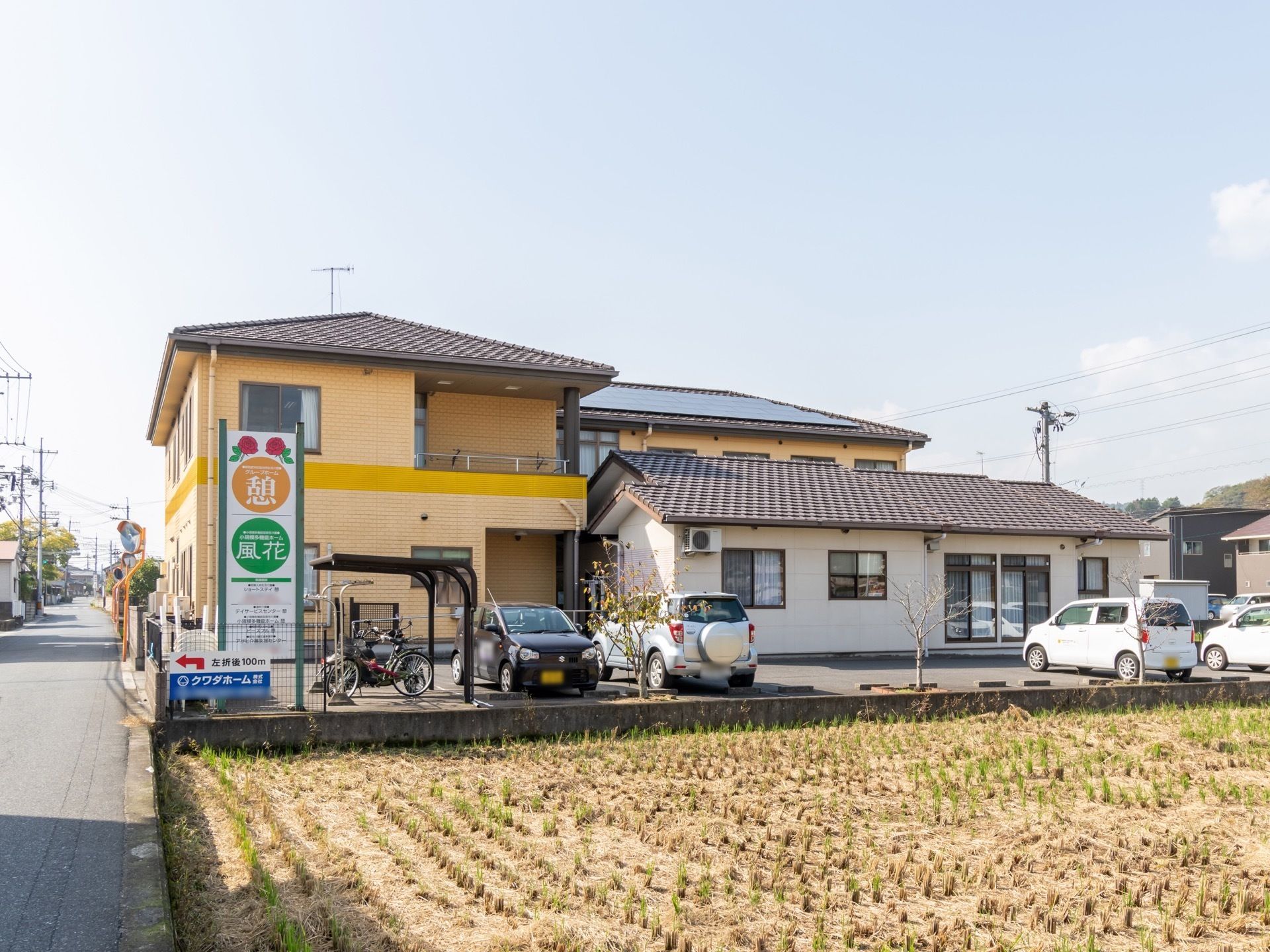 黄色の外壁の２階建ての大きな住宅風のホーム。敷地内に駐車場と駐輪場があり、敷地の端に看板も立てられている。