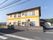 道路沿いにブロック塀に囲まれた、黄色の壁の２階建ての建物がある。建物の周囲には山や住宅、田んぼなどの景色が広がっている。