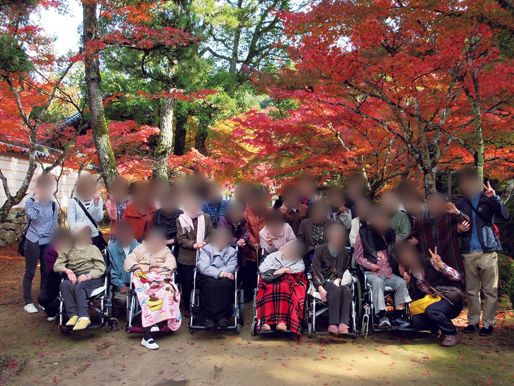 施設でのイベントの一環として、入居者様とスタッフで一泊旅行に出かけている。紅葉の木の前で、前後2列になって並んでいる。