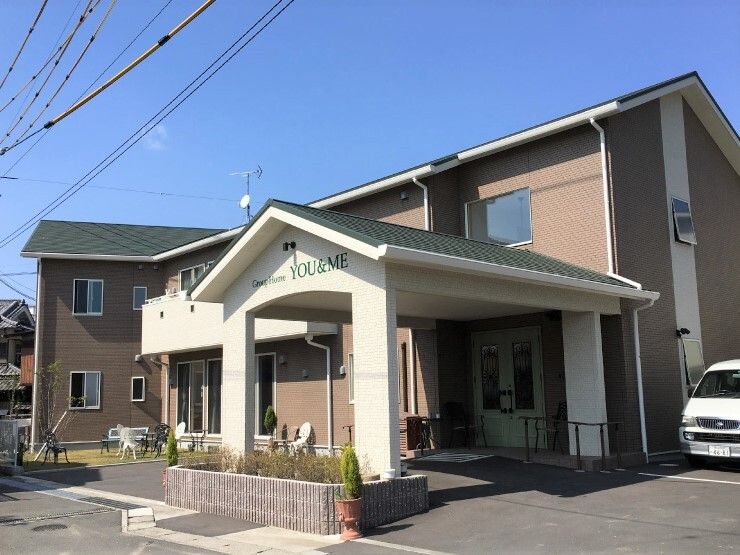 ブラウン系の2階建ての建物には屋根が付き、玄関部分には車寄せが付いている。敷地内には駐車場もあり、白いバンが止まっていて、庭があるのも見える。