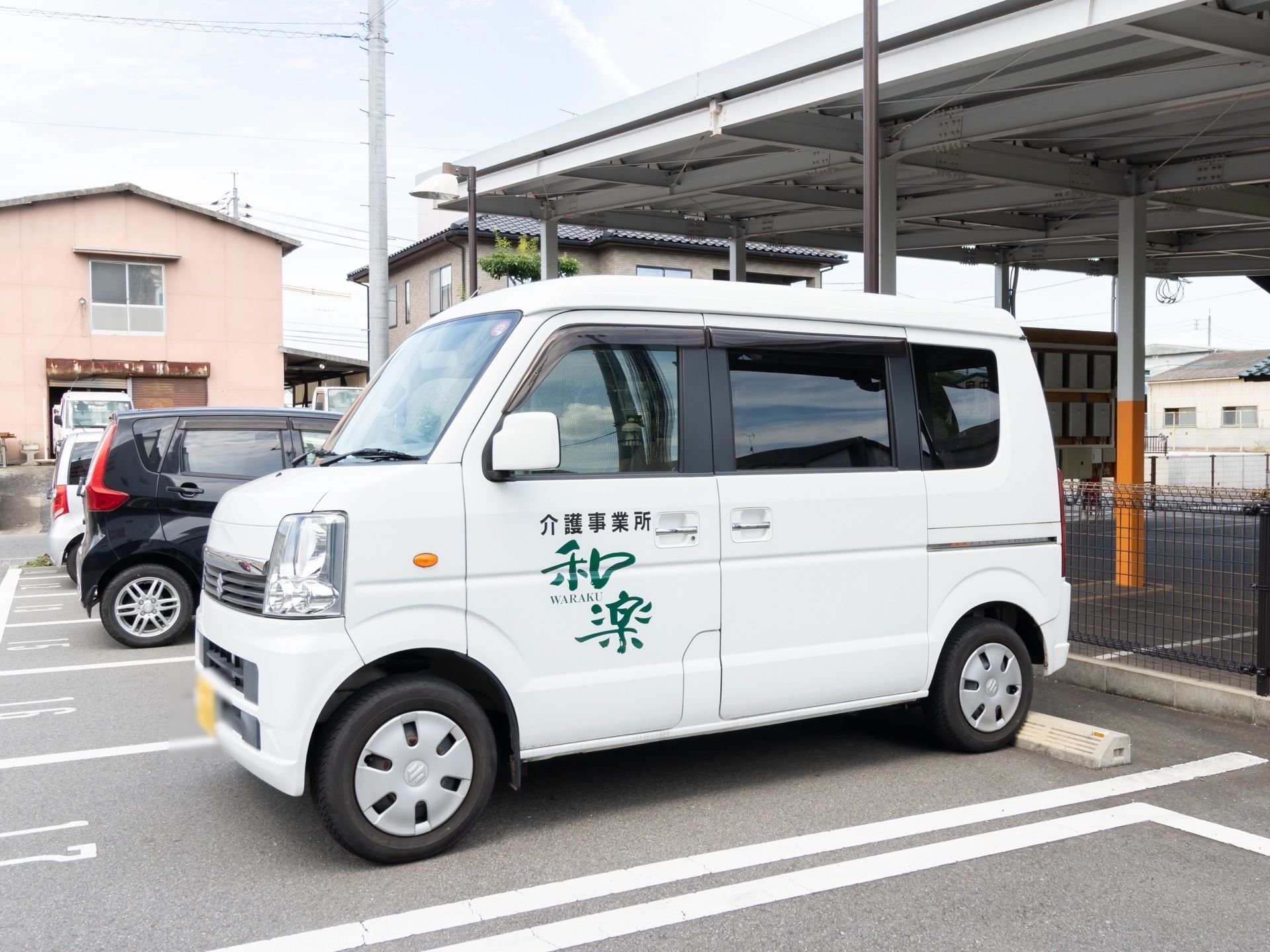 駐車場に施設名の書かれた白い車が停まっている。隣にも数台車が停められており、車の後ろにはフェンスが建てられている。