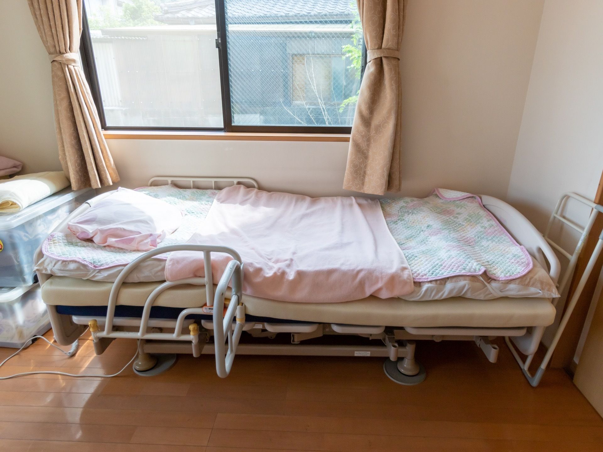 明るい光が差し込む窓の側に、介護ベッドがある。ベッドにはマットや枕などの寝具がセットされており、サイドレールがかかっている。