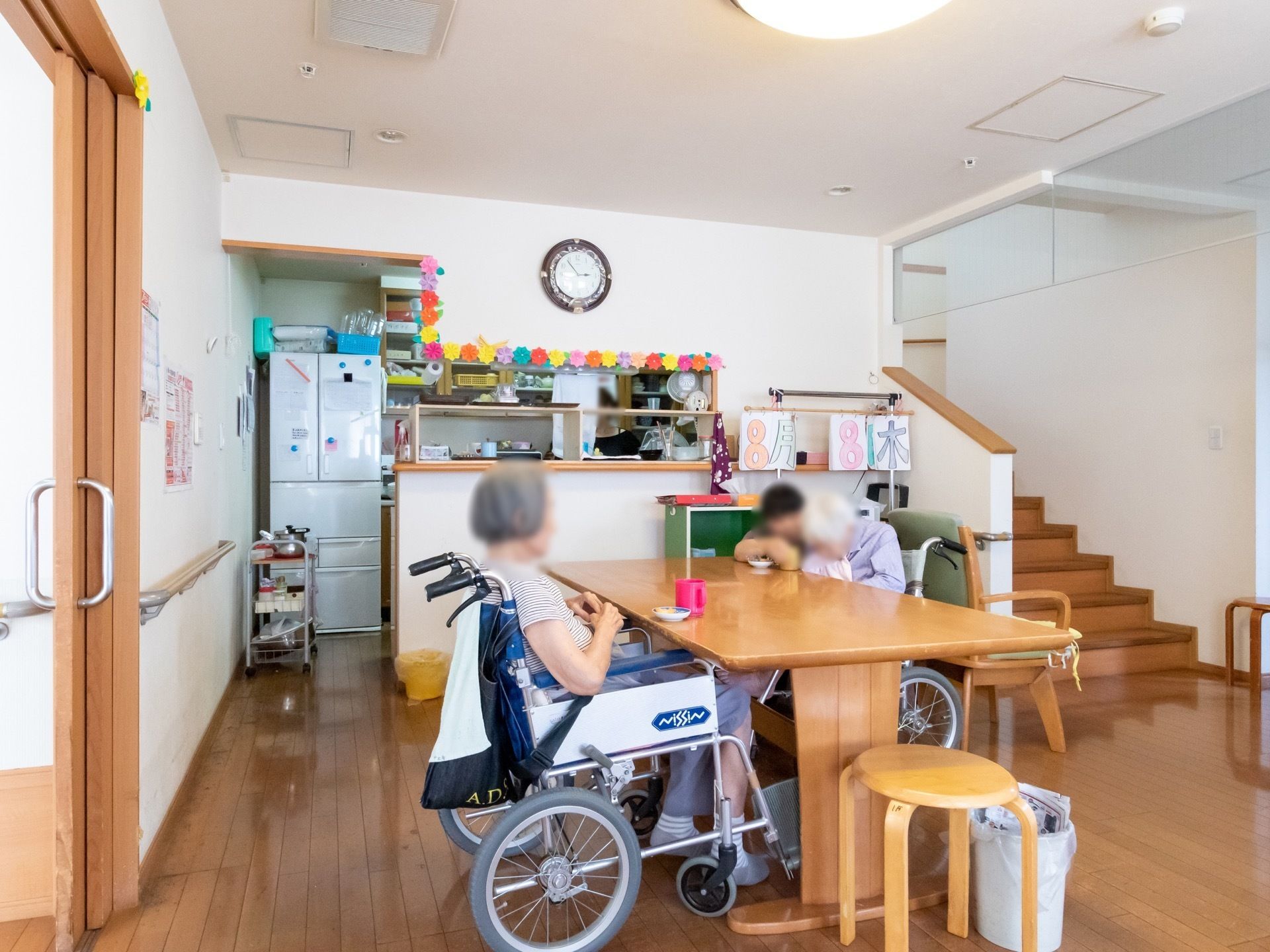 食堂の中央にはテーブルが設置されており、入居者様が2人座っている。奥にはキッチンがあり、冷蔵庫が置いてある。