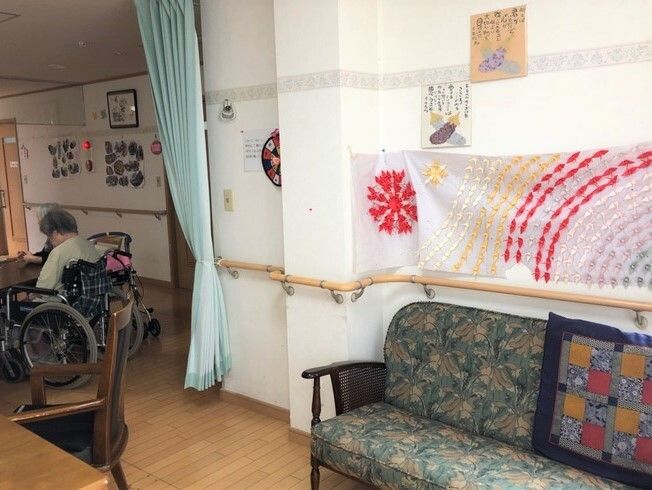 クッションが置かれたソファが置かれ、壁にはたくさんの入居者の作品が飾られ、奥には車いすの高齢女性がいる。