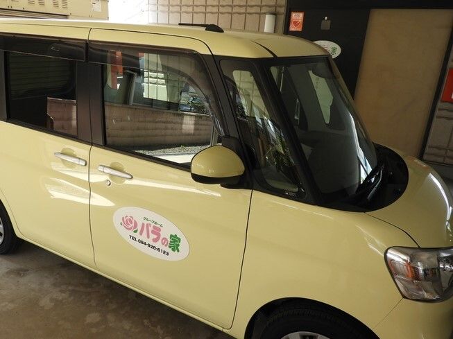 クリーム色の小さな車は建物の1階部分の駐車場に止められていて、周りは薄暗い。運転席のドアにホーム名が書かれている。