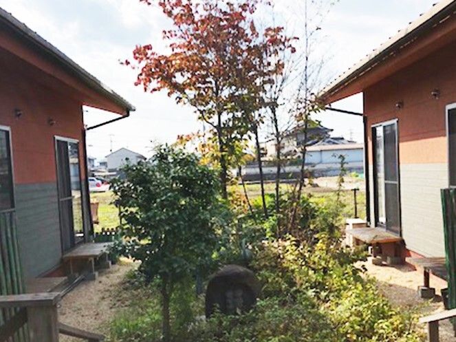 両サイドにオレンジの外壁の建物があり、それぞれの窓の掃き出し口にはベンチがある。庭には緑豊かな木が植えられている。