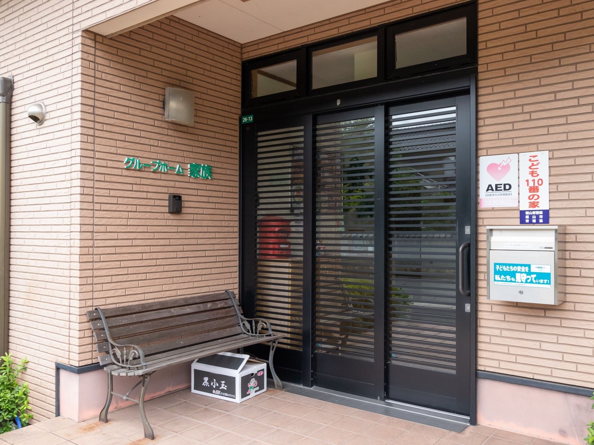 3枚スライド式扉が設置された施設入口である。木製のベンチが置かれ、ベージュ系のレンガ調外壁には、ポストがかかっている。