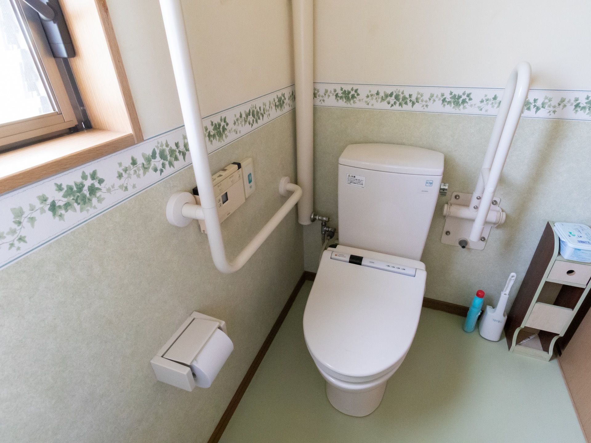 温水洗浄便座器が完備されたトイレである。デザイン性のある壁には、L字型や可動式手すりがある。