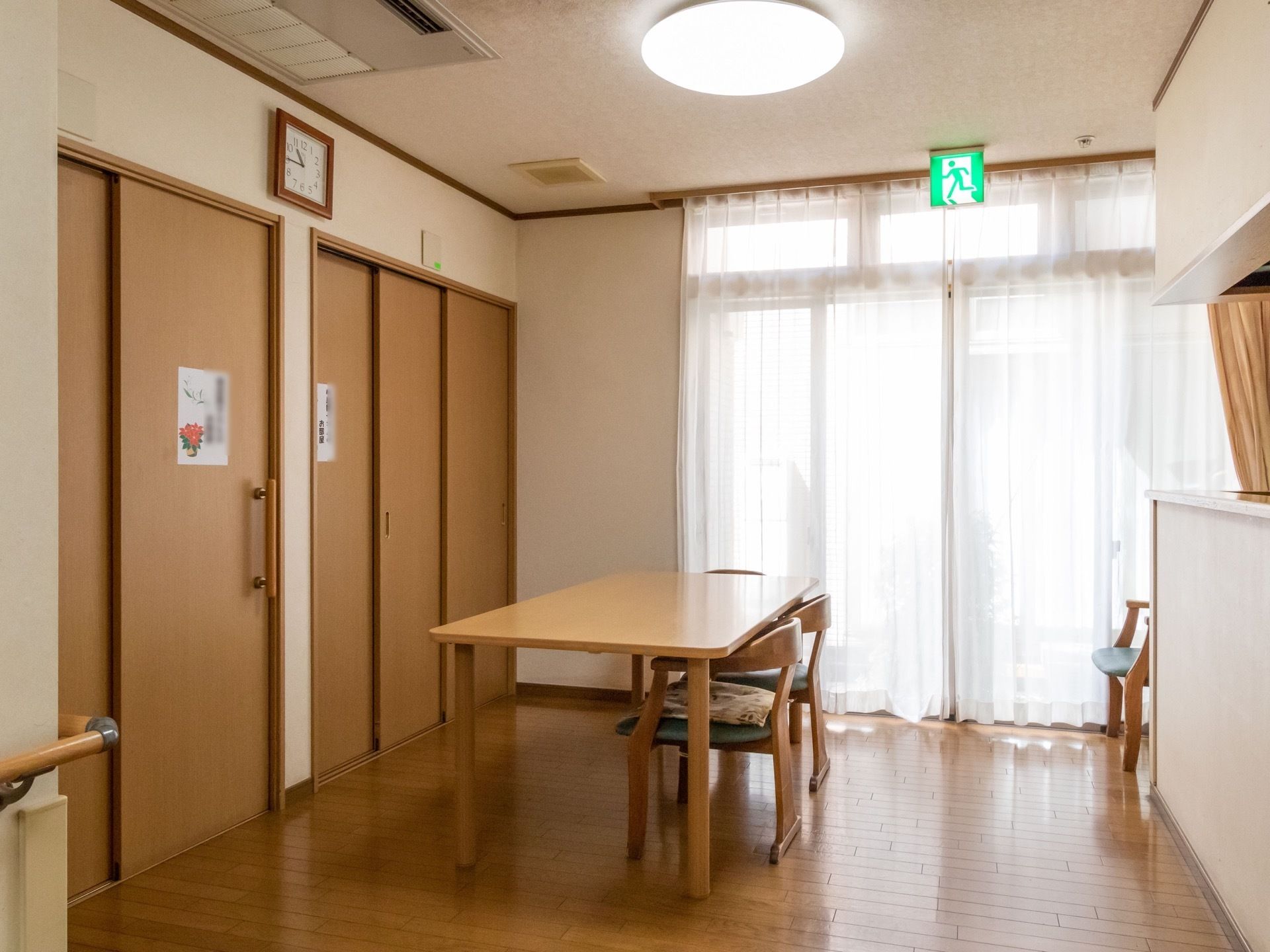共有スペースでは高さある窓が設置され、上部には非常口誘導灯がかかっている。明るい木目調のテーブルセットが配置されている。