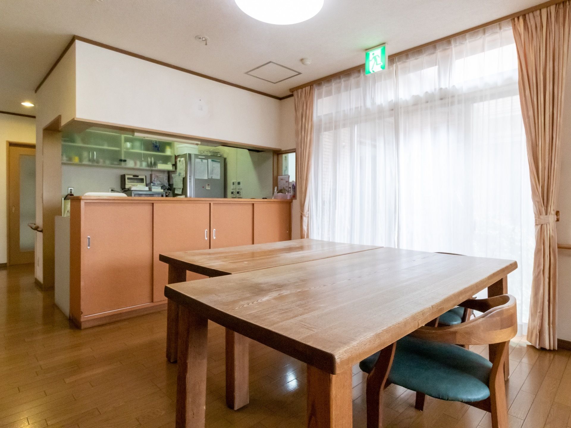 窓際の明るい場所に大きなダイニングテーブルがある。対面式のキッチンが併設され、スタッフが作業しながら入居者様の様子が確認できる。