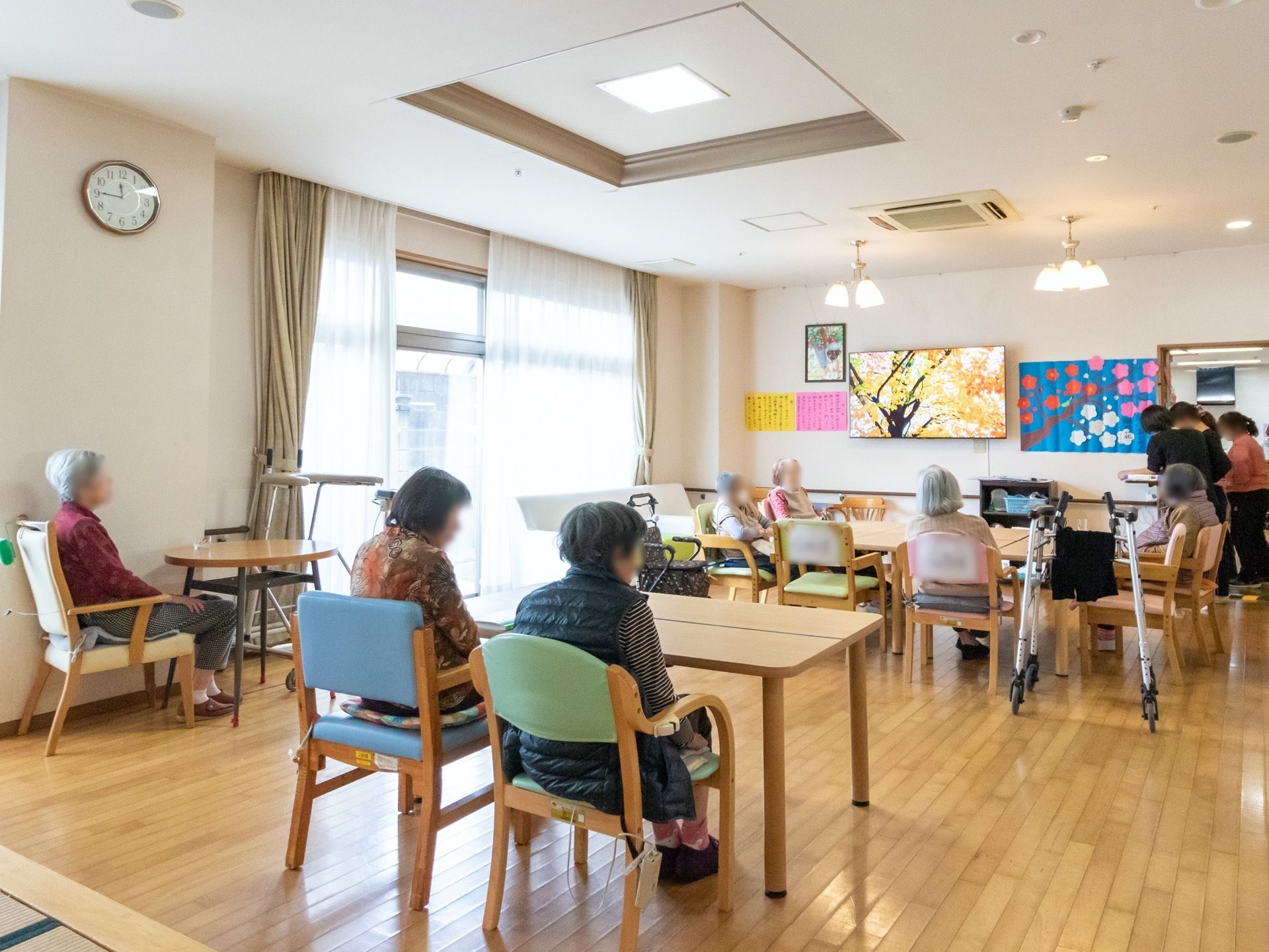 複数のテーブルが並べて置かれており、入居者様が座っている様子である。前方の壁には、カラフルな飾りが飾られている。