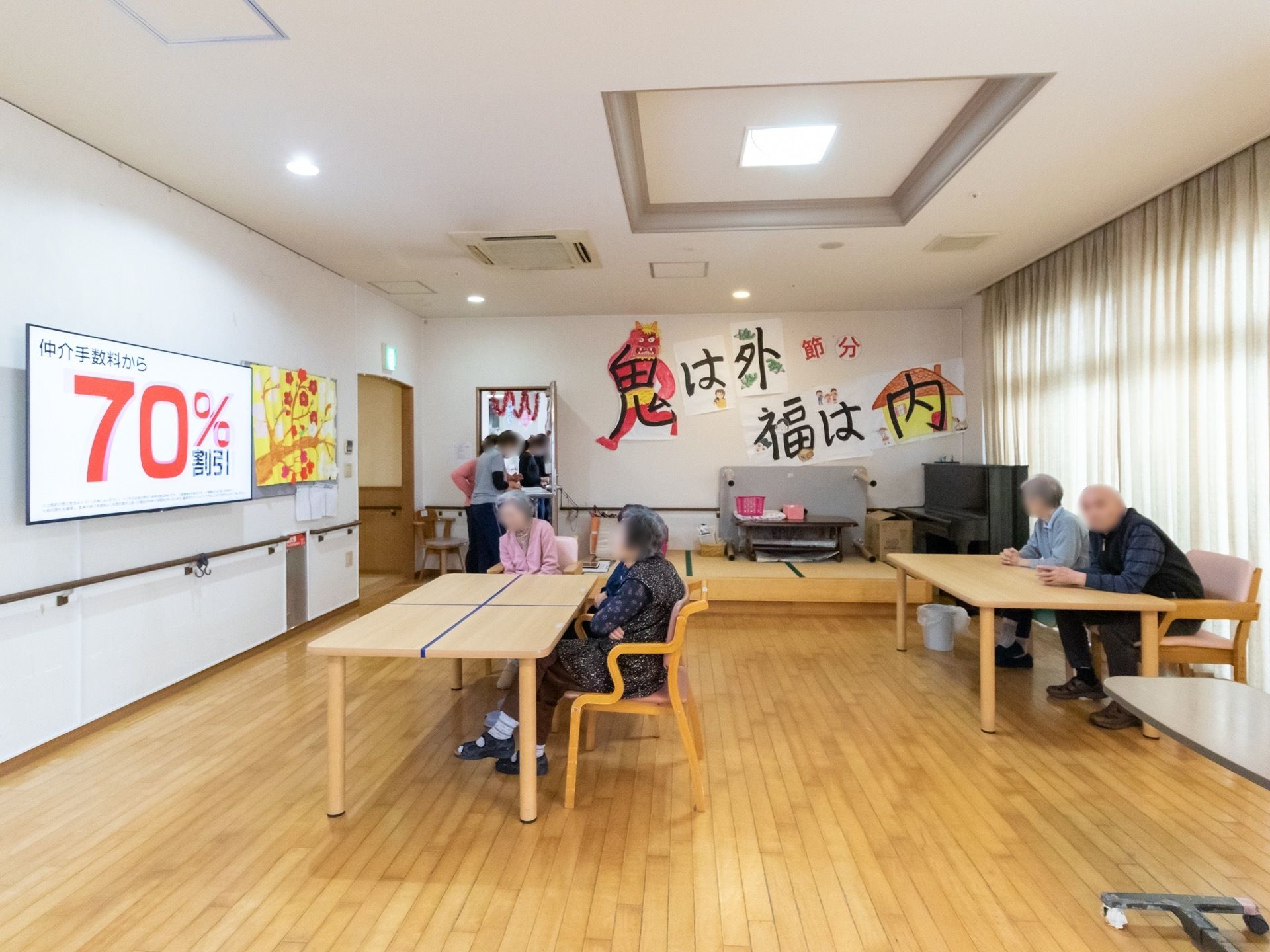 白い壁にかけられている、大きなテレビを見ている入居者様の様子である。壁には季節の飾りが飾られている。