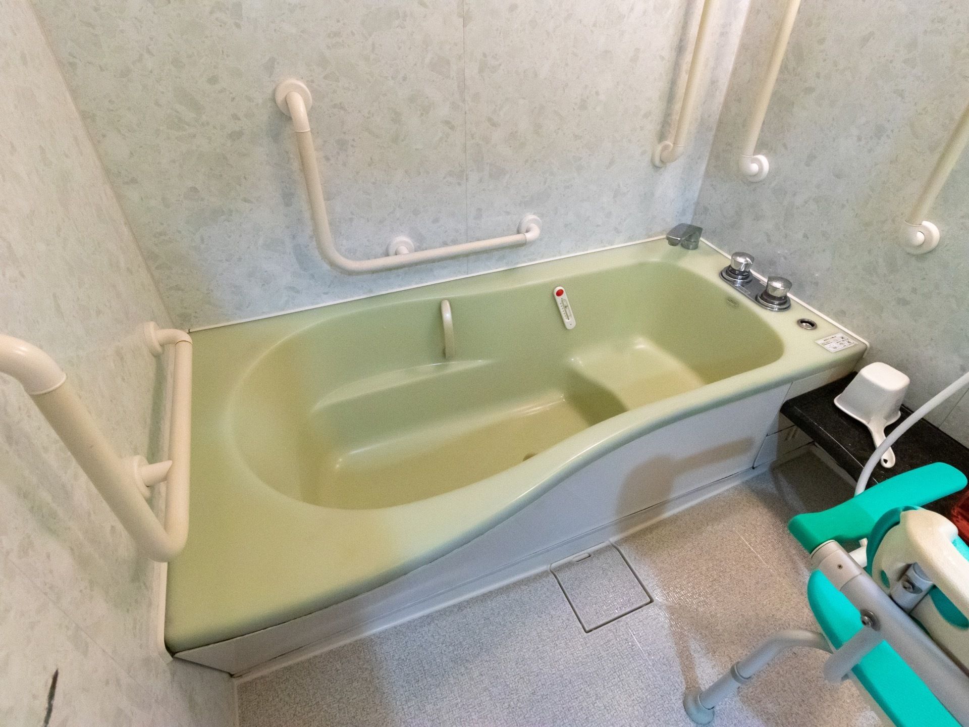 模様のある壁紙の浴室である。黄緑色の浴槽があり、壁には白色の手すりが壁に、複数本取り付けられている。浴室用チェアがある。