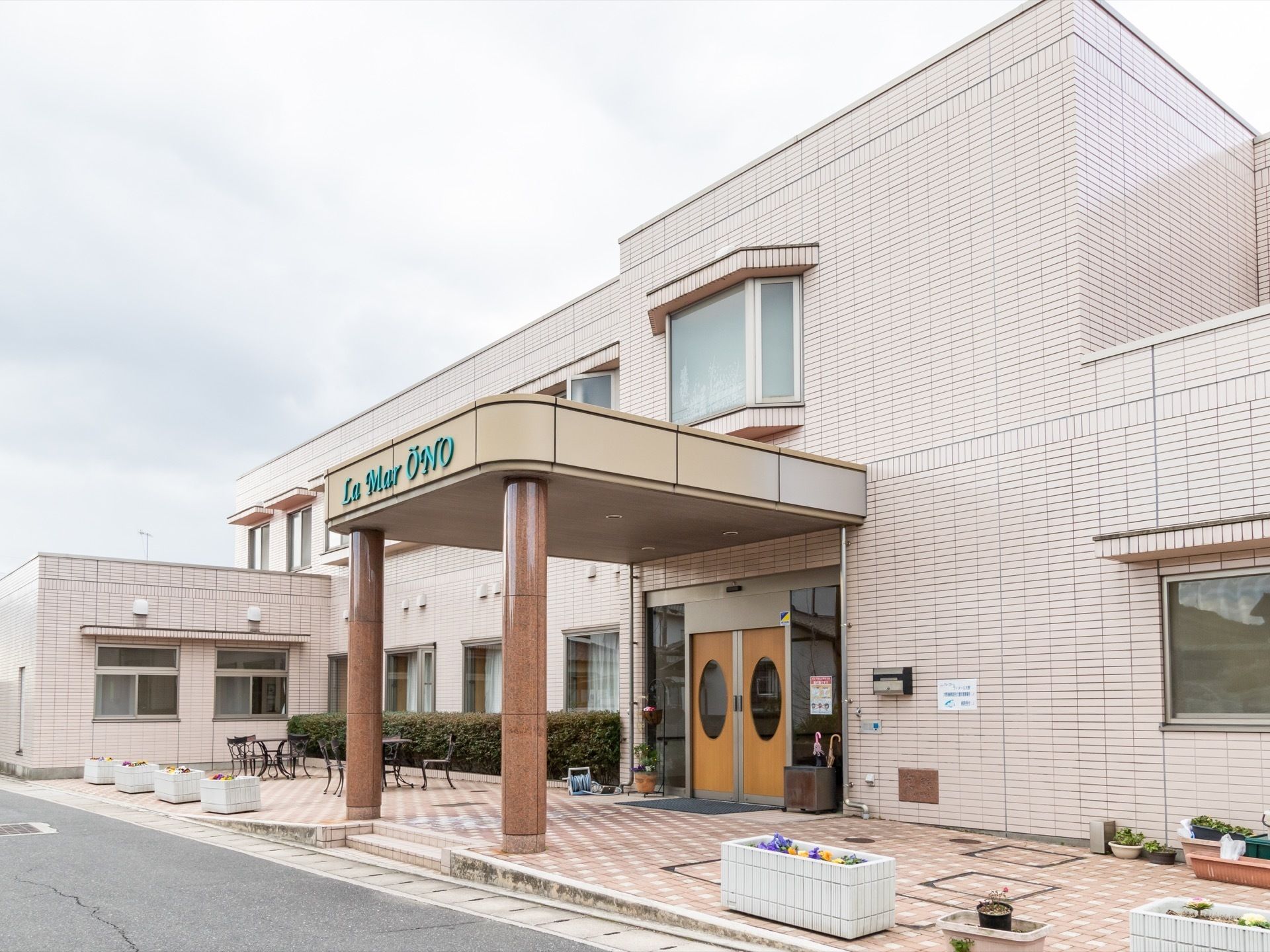 コンクリート造りの２階建ての建物である。２階には複数の出窓があり、１階には大きな窓がある。外にはテーブルと椅子がある。