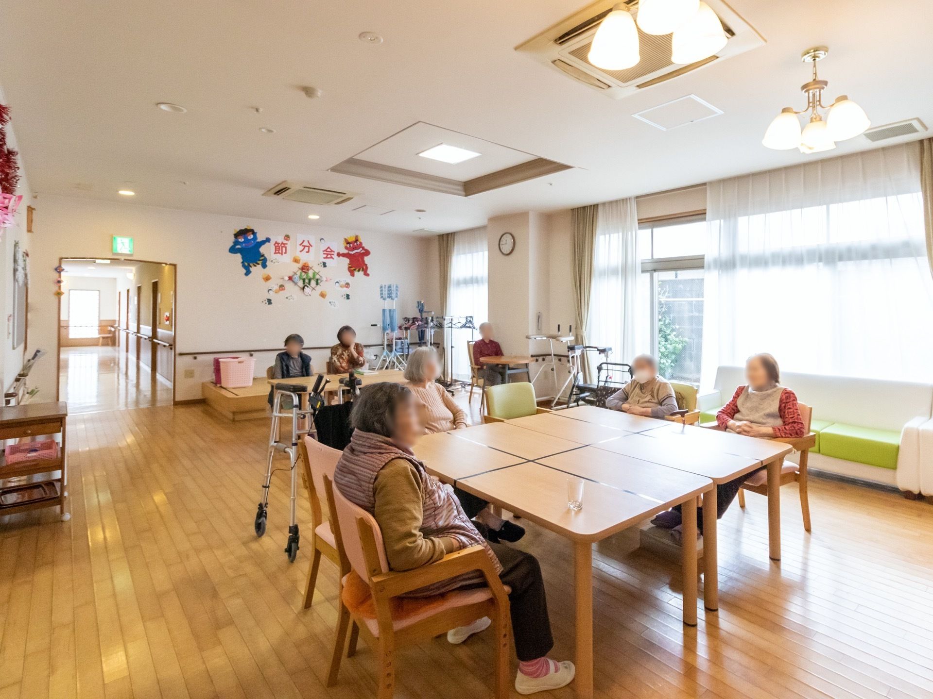 部屋の中央にテーブルを複数合わせて大きなテーブルを作り、その周りに入居者様が集まっている様子である。奥には小上がりがある。