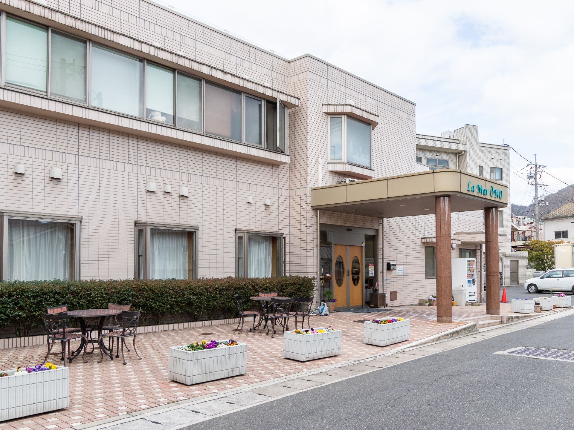庇付きの玄関がある建物の前にある、花壇とカフェテーブル、椅子