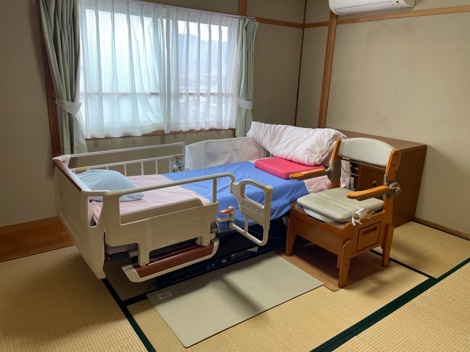 和室のお部屋。介護ベッドがあり、窓には水色の窓がある