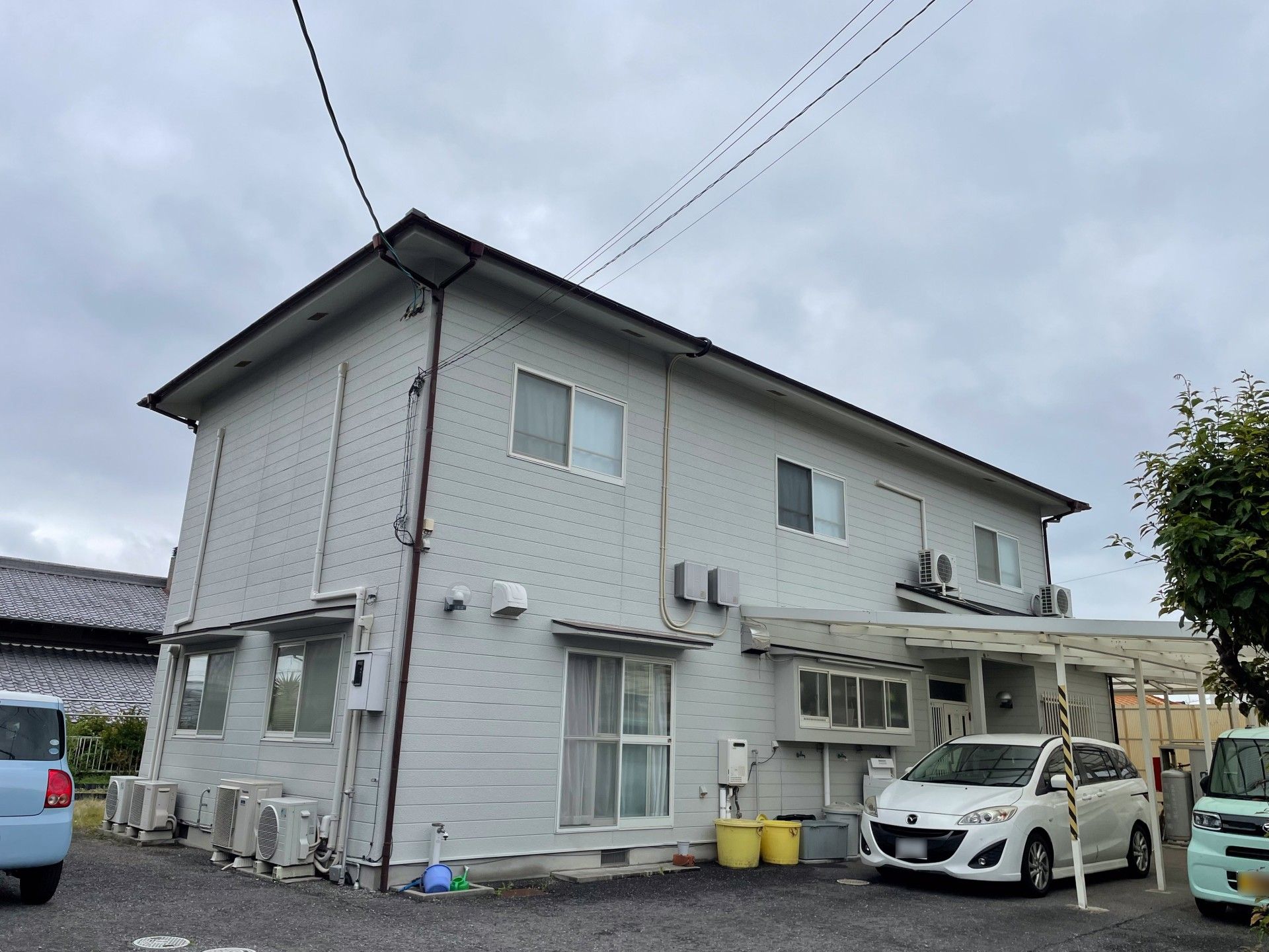 2階建ての一軒家。薄茶の外壁。緑色の車もあり
