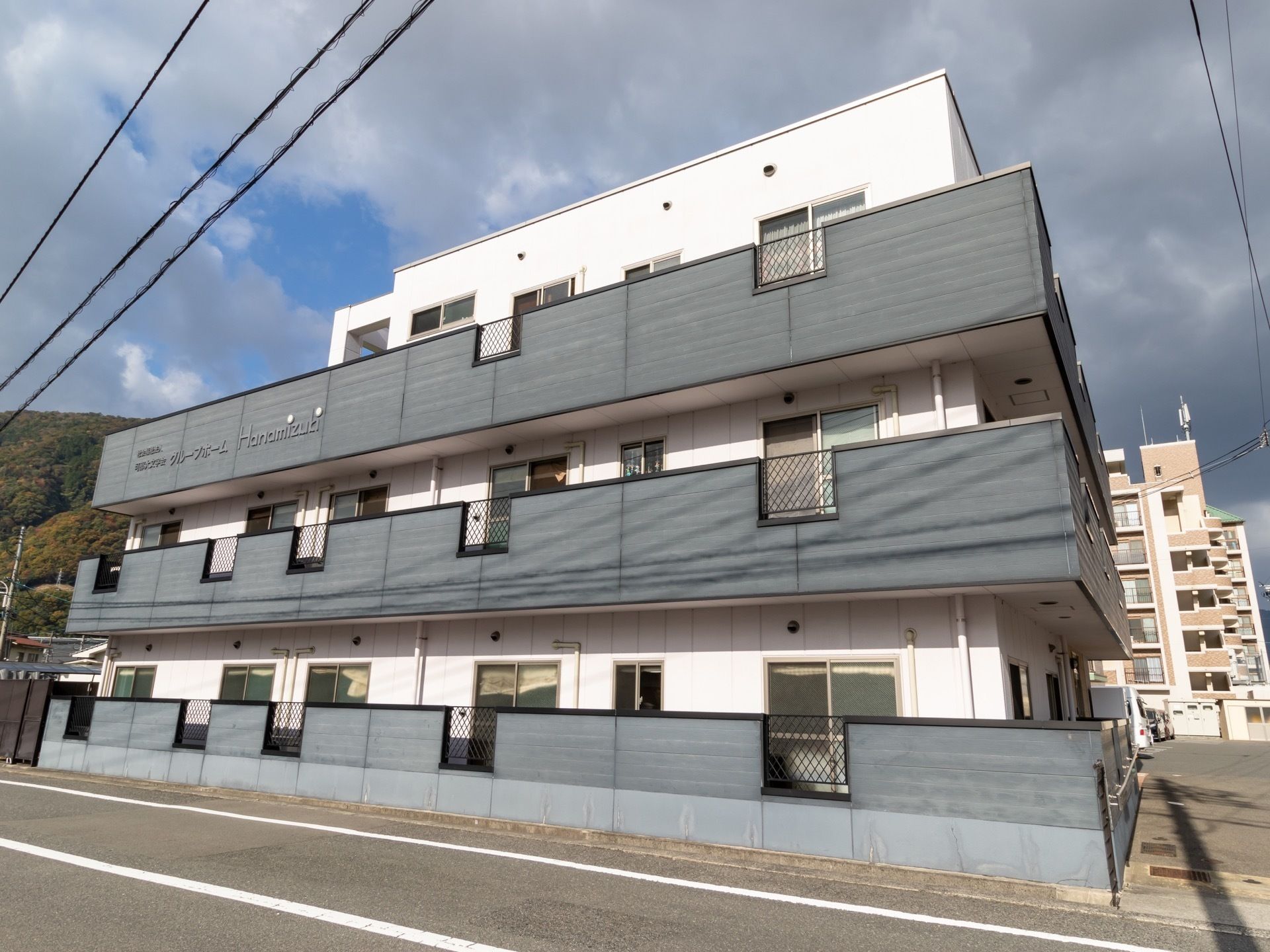 施設は３階建ての建物である。各階にダークグレー色の大きなベランダが設置されており、施設名が表示されている。