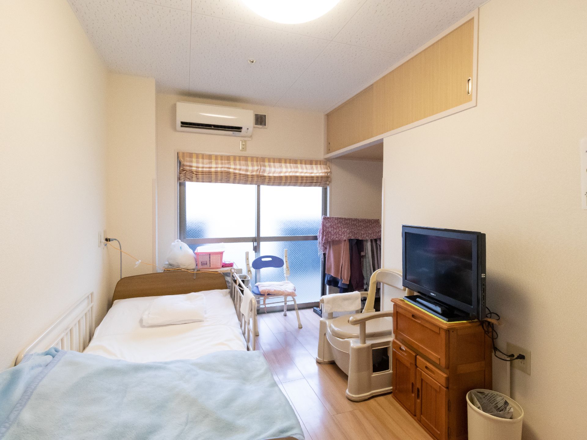 ベッド、テレビ、収納棚、ハンガーラックがあり、シェードカーテンが付いた居室