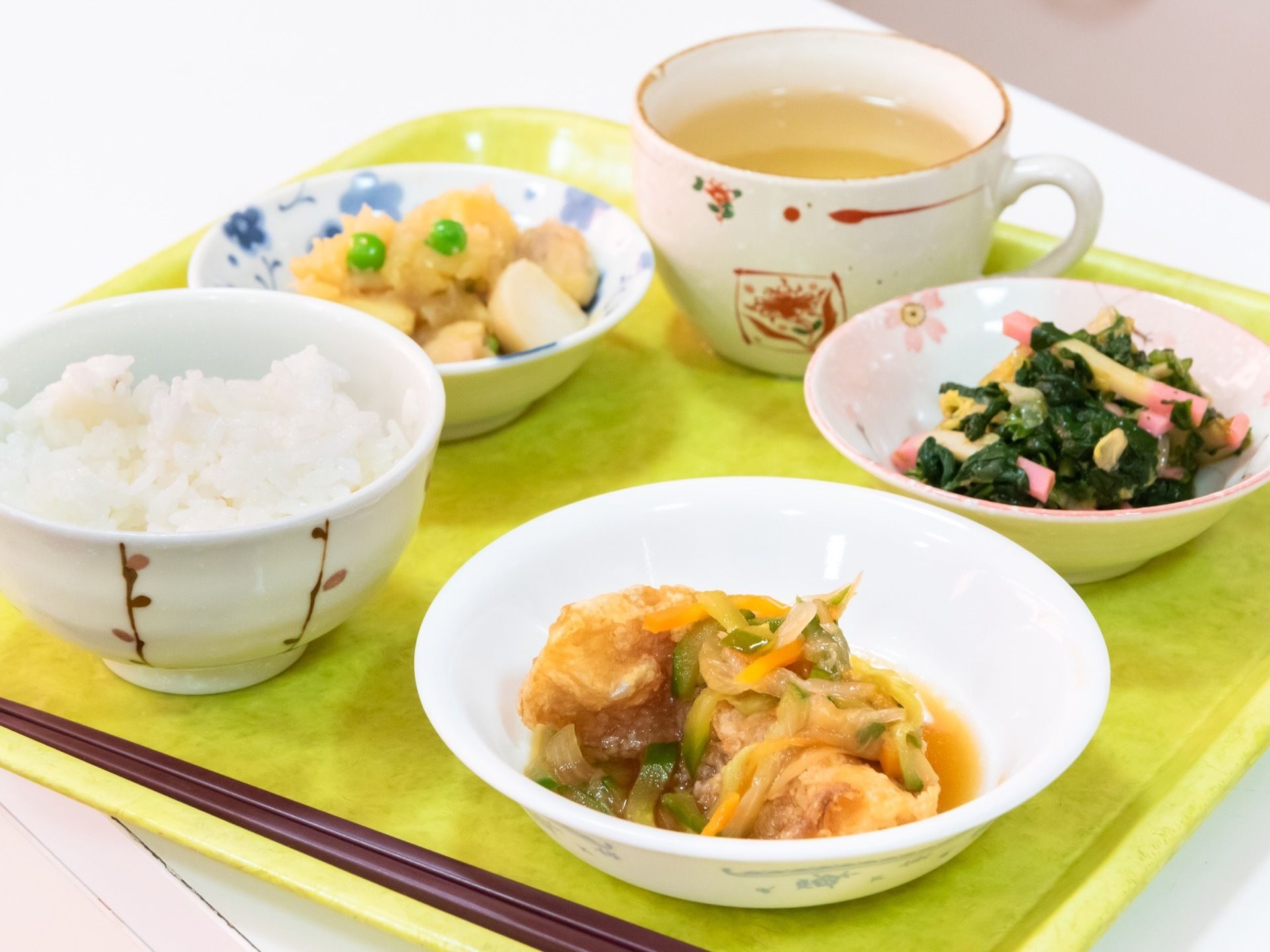 お茶と３種類のおかずとご飯が入った食器、箸などが黄色のトレイに載せられて、白い台の上に置かれている。