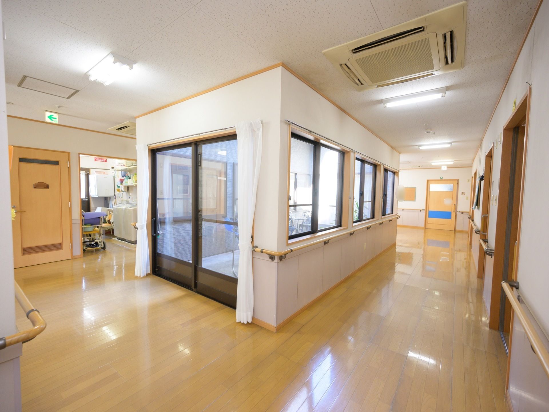 複数の窓がある明るい廊下である。天井には蛍光灯や空調設備が取り付けられており、壁には手すりがある。