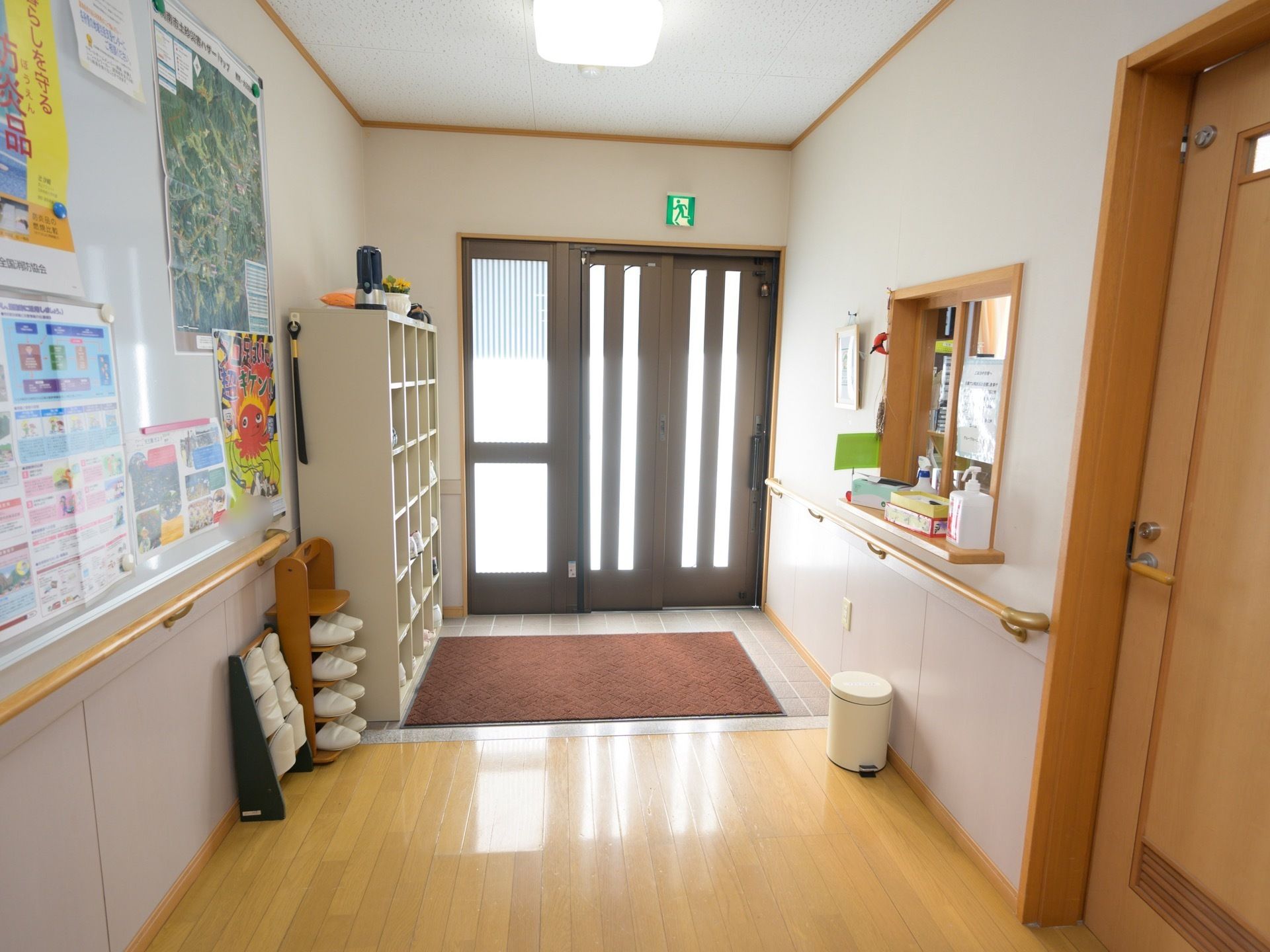 広々とした玄関で、事務室が併設されている。壁には手すりが設置されており、ホワイトボードには地図やポスターが貼られている。