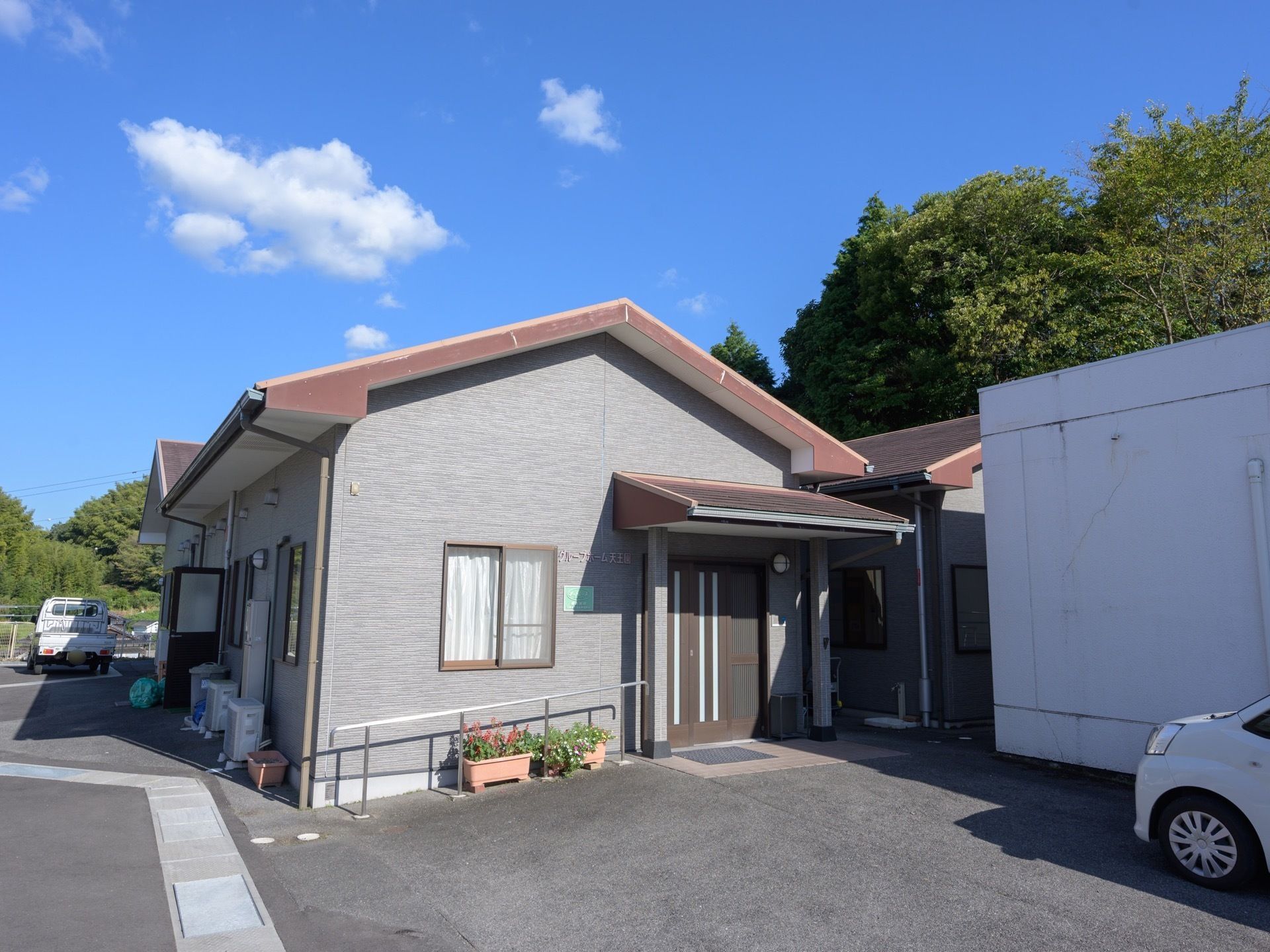 施設はグレーの外観の１階建てである。建物の奥に森が広がっている。建物の周囲に何台か車両を駐車することが可能。