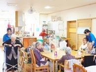 共有スペースに集まる入居者と介助に当たっているスタッフ。木製の家具が置かれ、家庭的な雰囲気。
